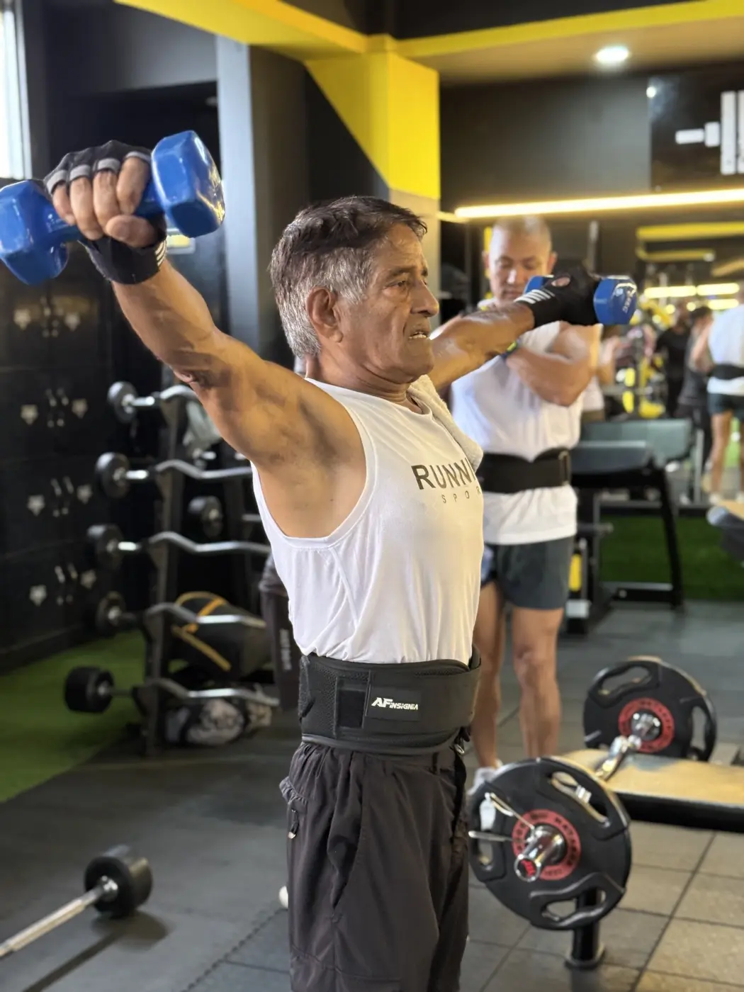 Older member pressing blue dumbbells with a lifting belt on